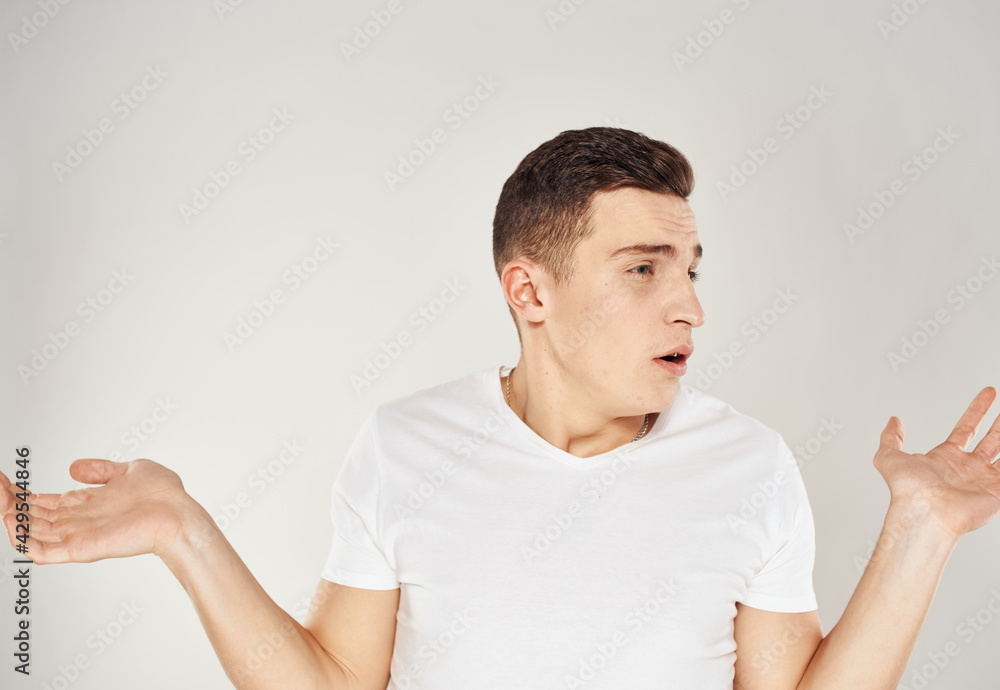 Fototapeta premium A man in bewilderment gestures with his hands on a light background in a white T-shirt cropped view