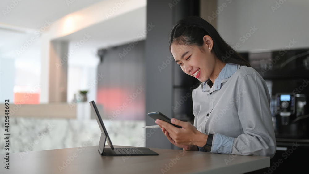 Cheerful young woman employee sitting in front of computer tablet at office desk and using smart phone. Cheerful young woman employee sitting in front of computer tablet at office desk and using smart phone.
