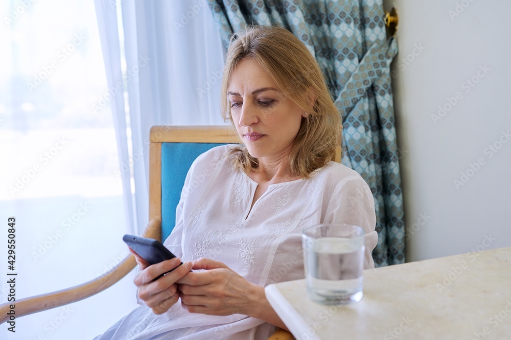 Serious middle aged woman with smartphone sitting on chair at home Serious middle aged woman with smartphone sitting on chair at home