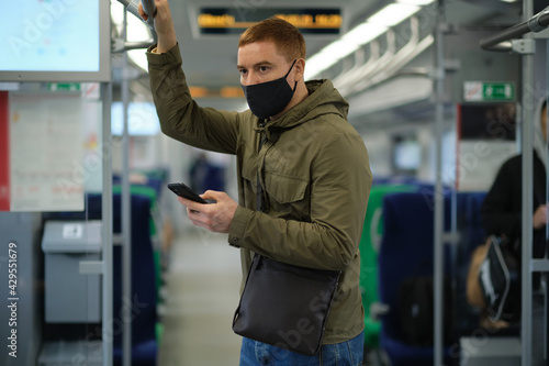 Handsome guy in respirator use a smartphone and keep social distancing to crowd while commuting in the metro or train. Infection and air pollution prevention concept