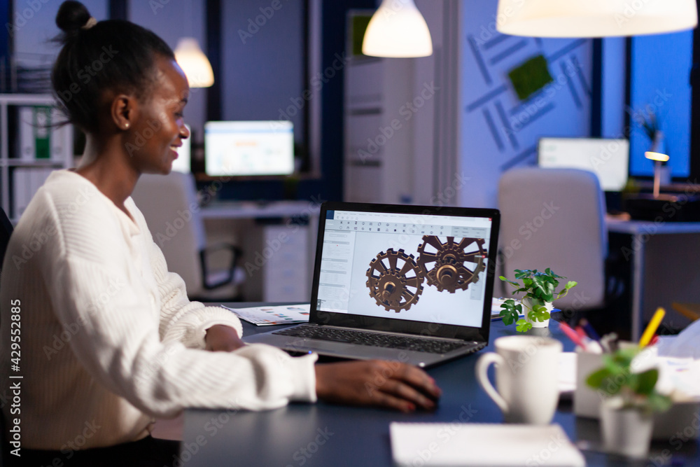 African engineer working late at night on a 3D model of industrial gears at laptop doing overtime in start up office. Freelancer studying prototype idea on pc showing cad software on device display.