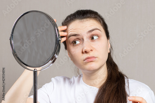 A young woman looks in the mirror and sees the first wrinkles. Anti-ageing facial skin care in cosmetology and dermatology.