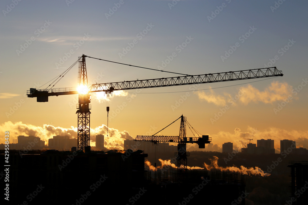 Silhouettes of construction cranes and unfinished residential buildings against the sky and shining sun. Housing construction, apartment block in city