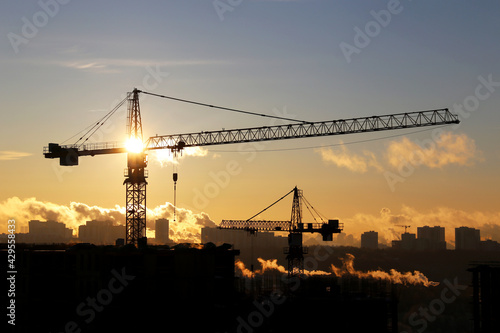 Silhouettes of construction cranes and unfinished residential buildings against the sky and shining sun. Housing construction, apartment block in city