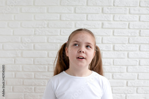 A girl in a white T-shirt looks away in surprise with her mouth open. White brick background. Copy space.