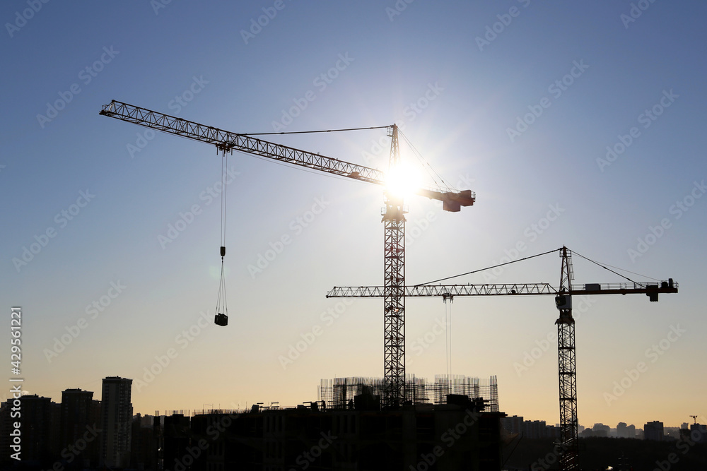 Silhouettes of tower cranes and unfinished residential building against the sky and shining sun. Housing construction, apartment block in city