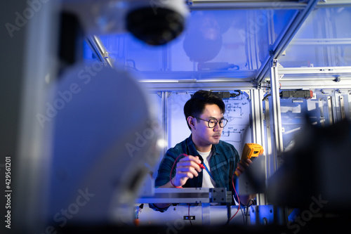Young Asian male electrical engineer looking at a digital multimeter's screen in hand while using it checking voltage to fix an industrial machine with a blurred of robotic machine in the foreground.