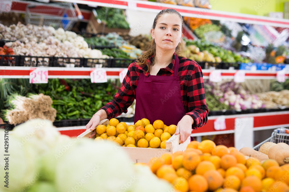Portrait of a young saleswoman standing with a box of tangerines near the counter in a store
