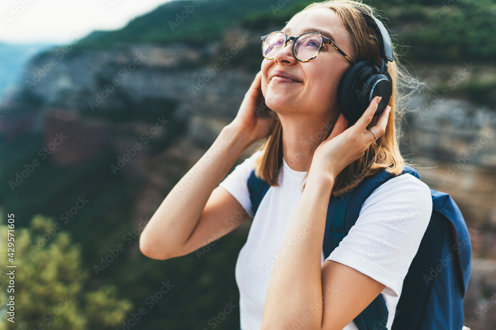 pretty fun young tourist woman with backpack and  hipster glasses smiling listens favorite music with headphones enjoying freedom of  hiking background natura mountain landscape on vacation outdoors