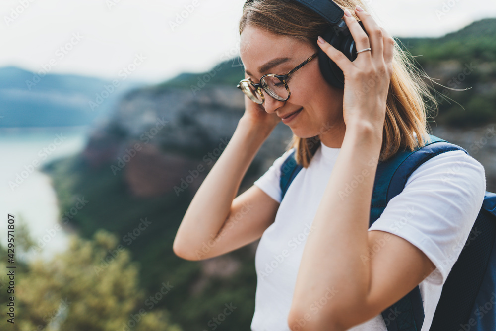 woman traveler with backpack and hipster glasses listening to favorite music on wireless headphones while hiking on top of mountain and enjoying relax freedom and fresh air outdoors