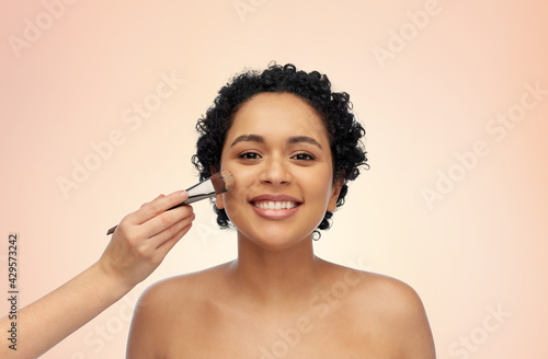 beauty, cosmetics and people concept - portrait of happy smiling young african american woman and hand of make up artist with brush applying foundation to her face over beige background