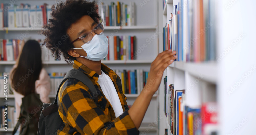 Young african-american man wearing mask looking for book in library Young african-american man wearing mask looking for book in library