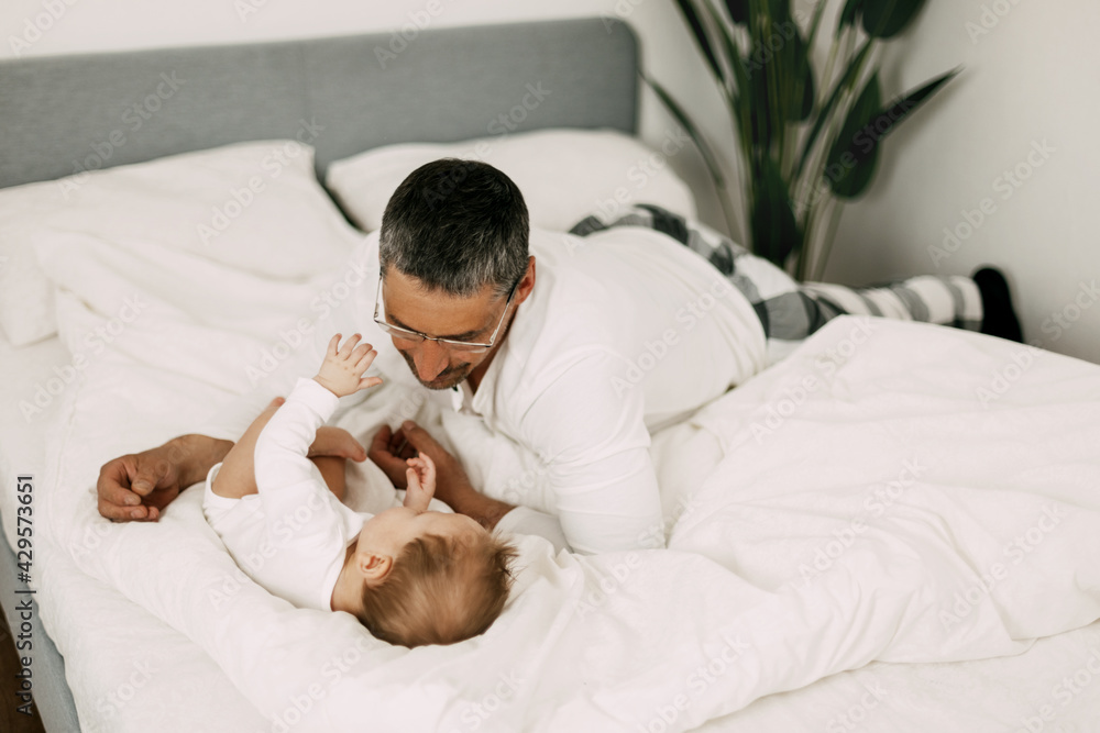 Happy father playing with adorable baby in the bedroom in a snow-white bed