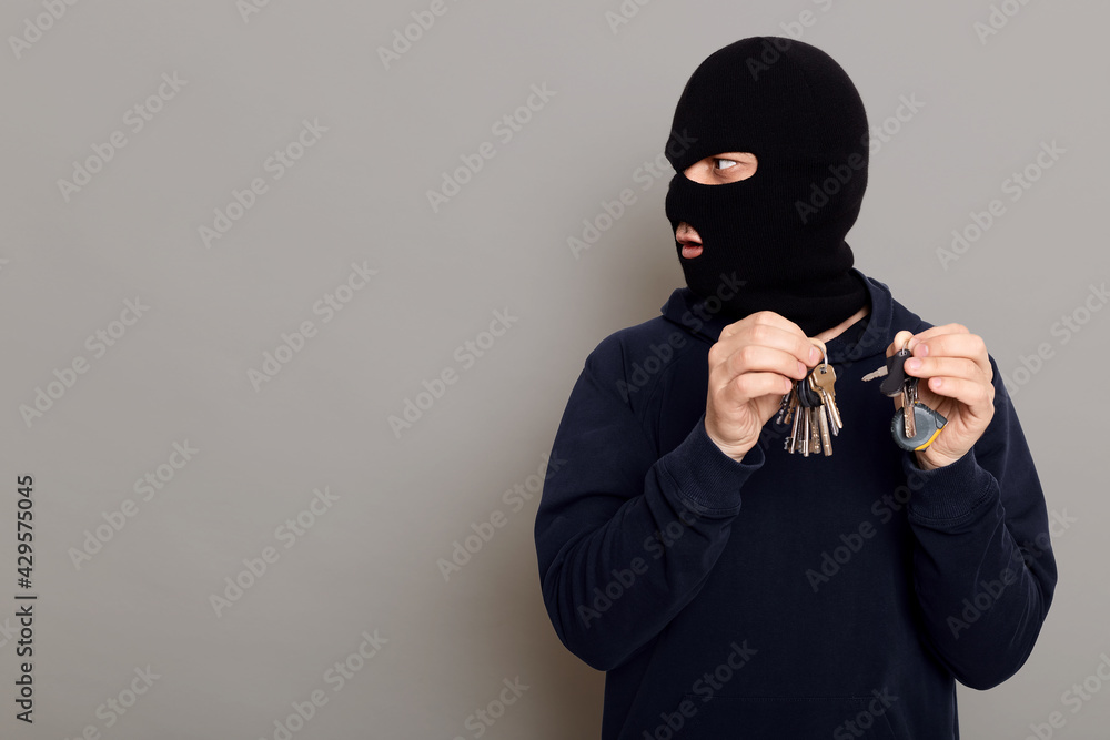 Surprised criminal thief looks to side and with contented expression on his face, holds in both hands bundle of keys, copy space for advertisement, posing isolated over gray background.