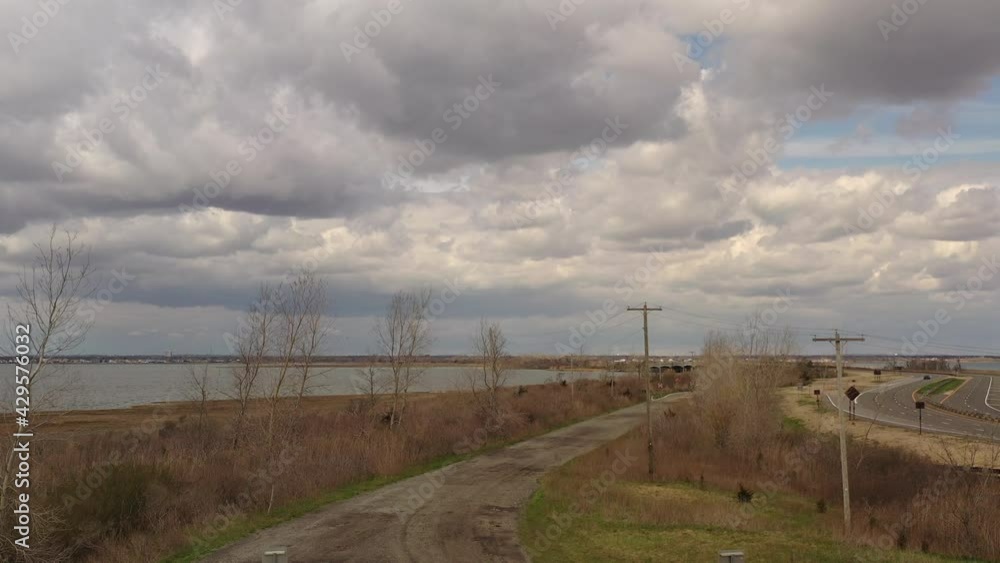 A low angle drone shot on a cloudy day, over a dirt road near a quiet bay. The drone camera slowly bloom up to reveal the waters and the cloudy sky. A low angle drone shot on a cloudy day, over a dirt road near a quiet bay. The drone camera slowly bloom up to reveal the waters and the cloudy sky.