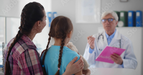 Aged male pediatrician sitting at table and writing prescription talking to mother and child