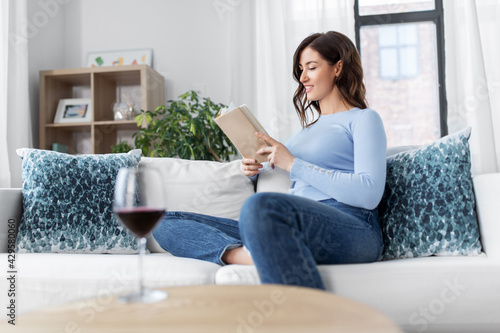 people and leisure concept - happy young woman sitting on sofa and reading book at home