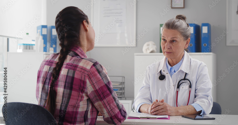 Obraz premium Senior doctor wearing white lab coat sitting at desk in office and talking to young woman patient.