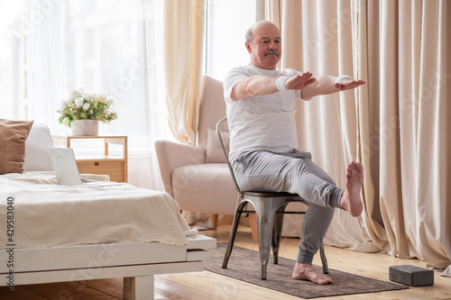 Elderly man practicing yoga asana or sport exercise for legs and hands on chair