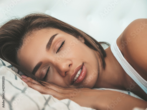 Portrait of young beautiful  female in white lingerie. Sexy carefree woman lies on bed in posh apartment or hotel room. She wakes up. She sleeping at sunny lazy morning. Closed eyes