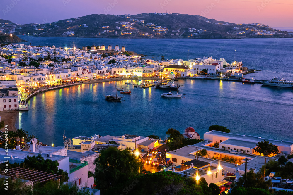 Colorful view after sunset of Mykonos, Greece, harbor and port, ships, whitewashed houses. Town lights up. Vacations, leisure, Mediterranean lifestyle