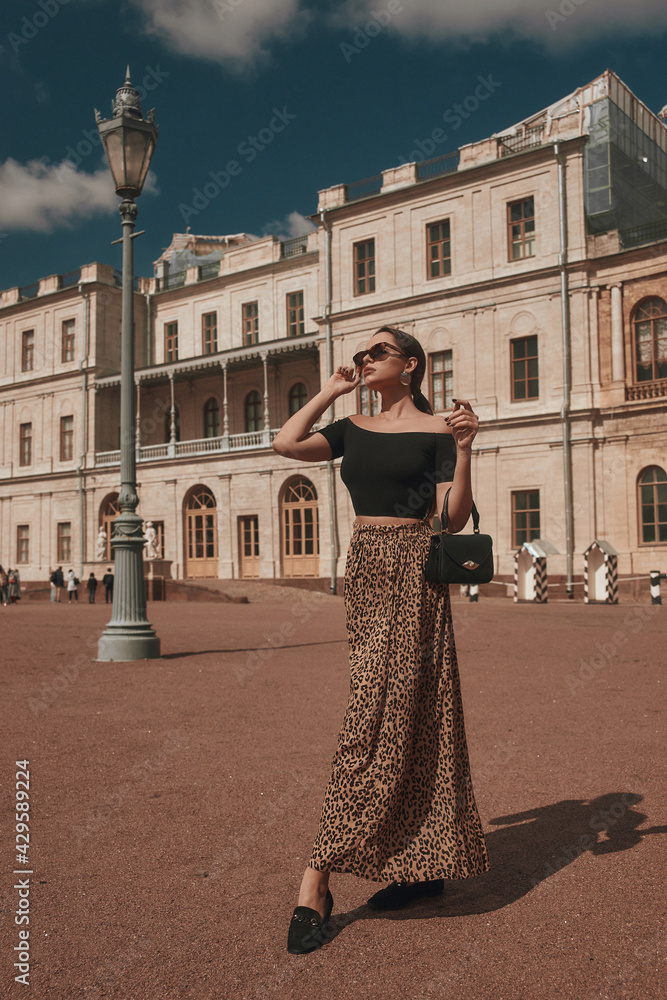 Elegant woman wearing black top, leopard skirt and sunglasses, holding black leather handbag, standing and posing at square against beautiful palace.