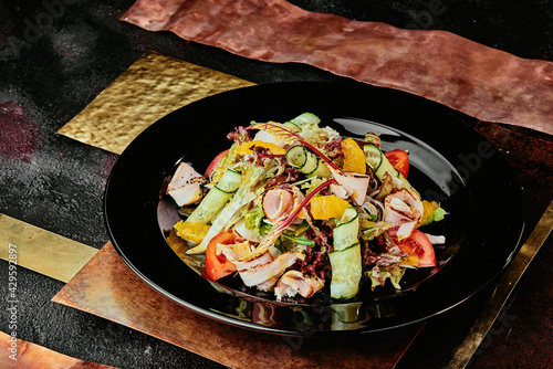 salad with ham, orange slices, tomato, cucumber and lettuce leaves in a black plate on a copper background. 