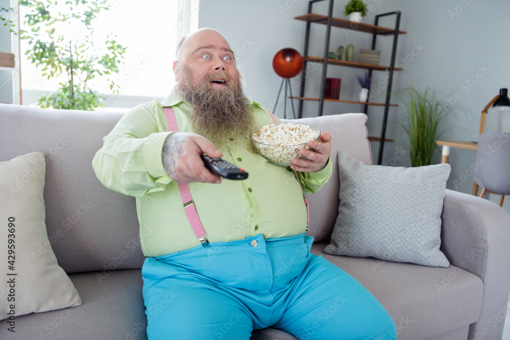 © deagreez - Portrait of impressed positive man sit on sofa hold remote control popcorn look interested tv weekend indoors