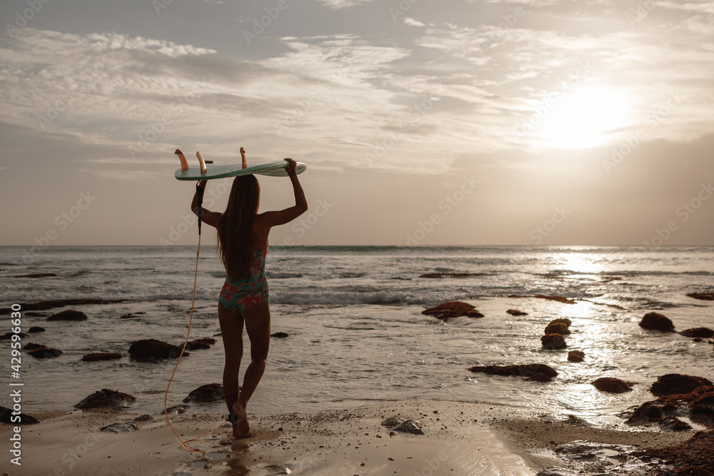  Attractive young woman in surf swimsuit is standing on beach with blue surfboard in hands over her head, look at sunse and go to the ocean catch waves