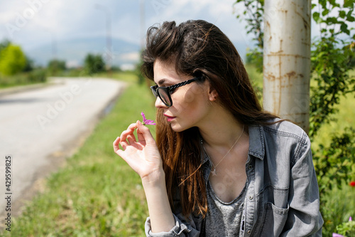 Young woman is sitting alongside the road and is smelling a flower.