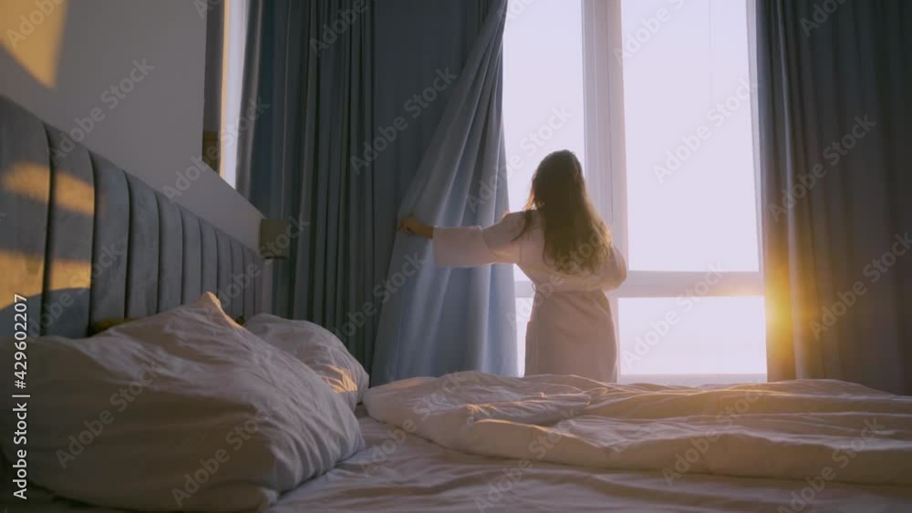 Young curly haired woman in white bathrobe opens curtains on large panoramic window looking outside in elegant hotel room at sunrise backside view