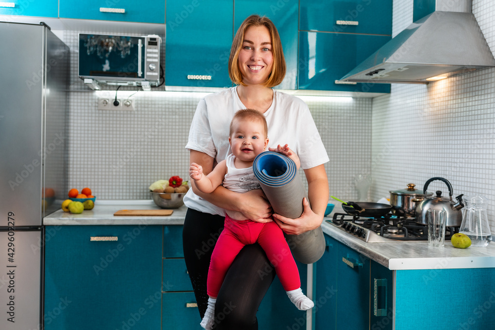 Portrait of a young happy mother holds her baby in her arms, and a sports mat rolled up. In the background is the kitchen. The concept of sports activity at home