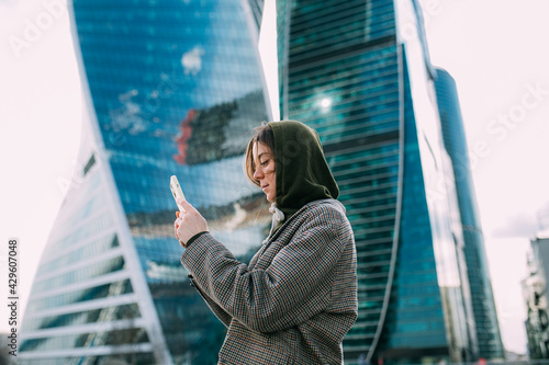 A blonde young girl dressed in a coat uses a smartphone against the backdrop of skyscrapers.Writes a message and calls. Side view. Modern technologies and communications photo