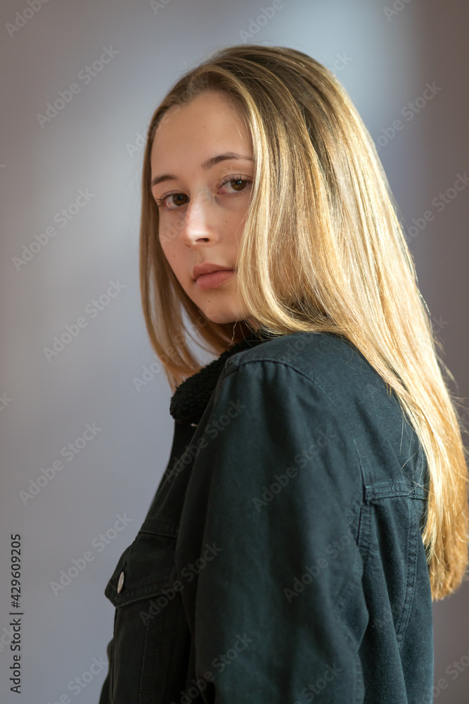 Fototapeta premium Three quarters shot or semi profile portrait of a pretty teenage girl with long blond hair posing looking at the camera over a grey background