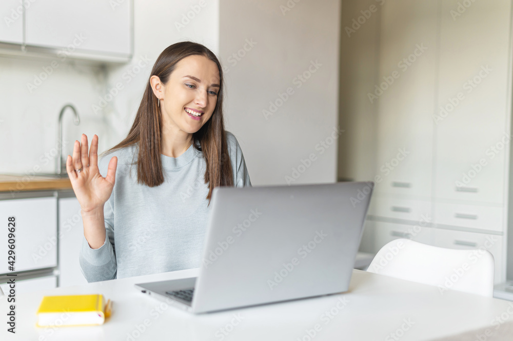 Video call concept. Happy young woman using laptop computer for video connection from home, female freelancer looks at webcam and greeting colleagues, waving and smiles sitting in the kitchen home Video call concept. Happy young woman using laptop computer for video connection from home, female freelancer looks at webcam and greeting colleagues, waving and smiles sitting in the kitchen home