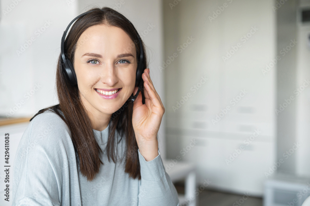 Headshot of smiling young female employee wearing wireless headset, looks at camera, cheerful woman in headphones takes a call from colleagues or clients online, working remotely from home office Headshot of smiling young female employee wearing wireless headset, looks at camera, cheerful woman in headphones takes a call from colleagues or clients online, working remotely from home office