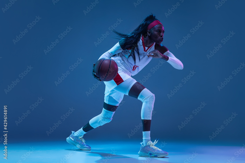 Obraz premium Beautiful african-american female basketball player in motion and action in neon light on blue background. Concept of healthy lifestyle, professional sport, hobby.