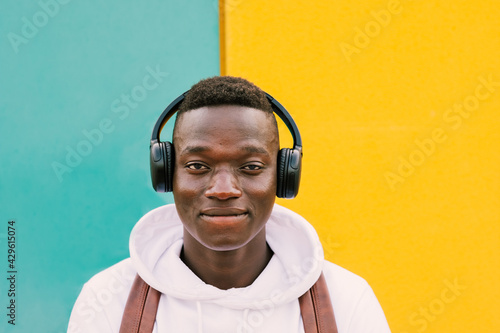 Young afro American black man listening music with wireless headphones while wearing a white sweatshirt and a backpack looking to camera on a green and yellow wall background