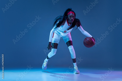 Beautiful african-american female basketball player in motion and action in neon light on blue background. Concept of healthy lifestyle, professional sport, hobby.