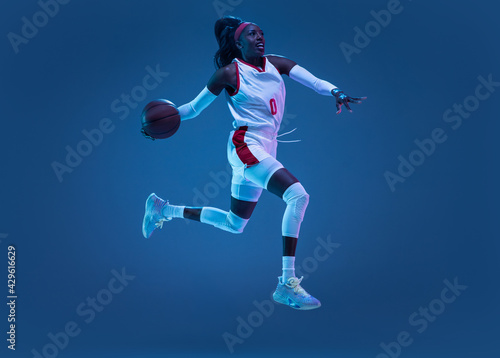 Beautiful african-american female basketball player in motion and action in neon light on blue background. Concept of healthy lifestyle, professional sport, hobby.