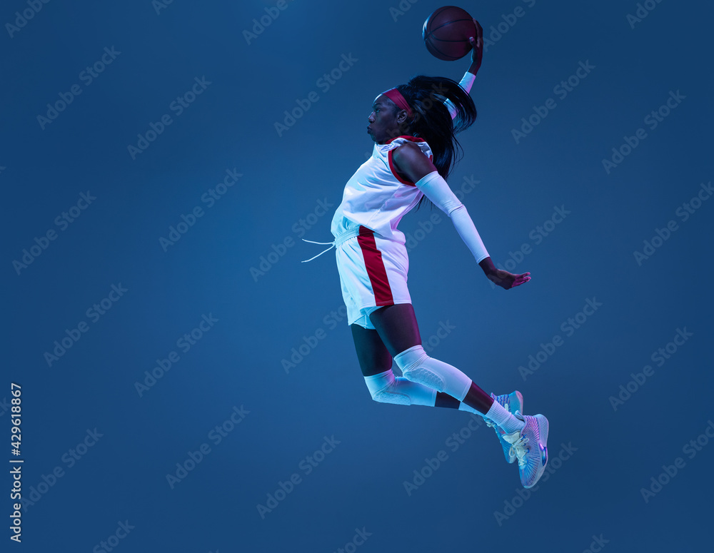 Beautiful african-american female basketball player in motion and action in neon light on blue background. Concept of healthy lifestyle, professional sport, hobby.
