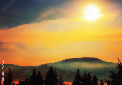 A colorful photo of a mountain landscape at sunset. forest, mountains and bright sun in red clouds © Ann Stryzhekin