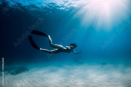 Free diver woman with fins glides underwater over sand in blue ocean.