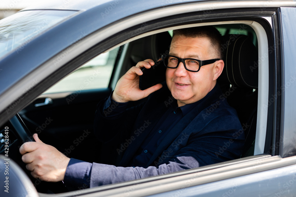 Cheerful man wearing eyeglasses sitting on the drivers seat and talking with his wife on the phone.
