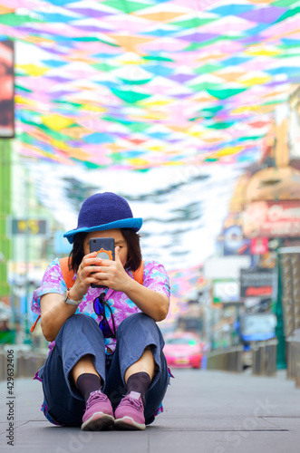 Asian female tourist use smartphone to taking picture while sitting in the middle of walking street with colorful decorative fabric hanging on the air in vertical frame