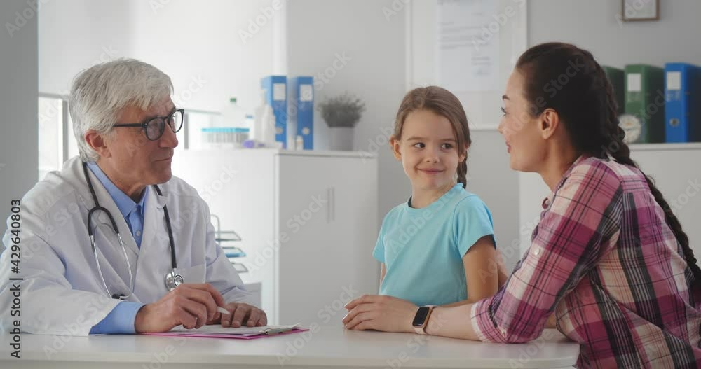 Aged male pediatrician doctor talking to little girl and mother in office of medical clinic. Aged male pediatrician doctor talking to little girl and mother in office of medical clinic.