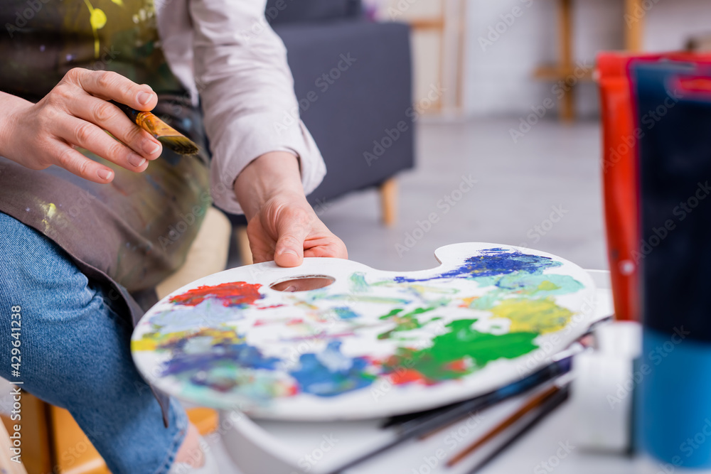 cropped view of middle aged artist holding paintbrush and reaching palette with colorful paints cropped view of middle aged artist holding paintbrush and reaching palette with colorful paints