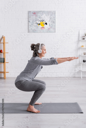 Bild auf Leinwand side view of cheerful middle aged woman in sportwear and headphones doing squat