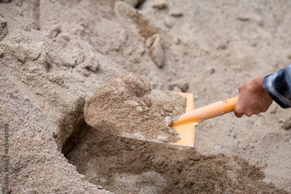 A close-up of the sand in the shovel, the hand of a construction worker shoveling the sand with a shovel. From sandpiles to mix cement on the construction site - obrazy, fototapety, plakaty 