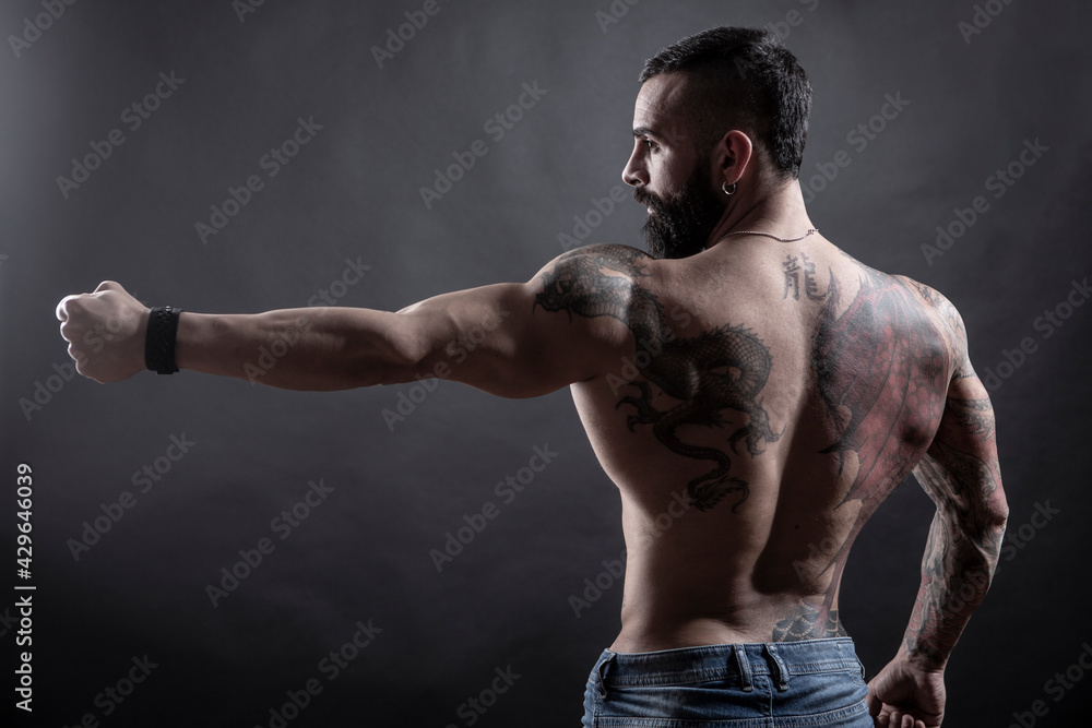 Uomo moro muscoloso e tatuato fa una posa mostrando il braccio e la schiena , isolato su sfondo nero
